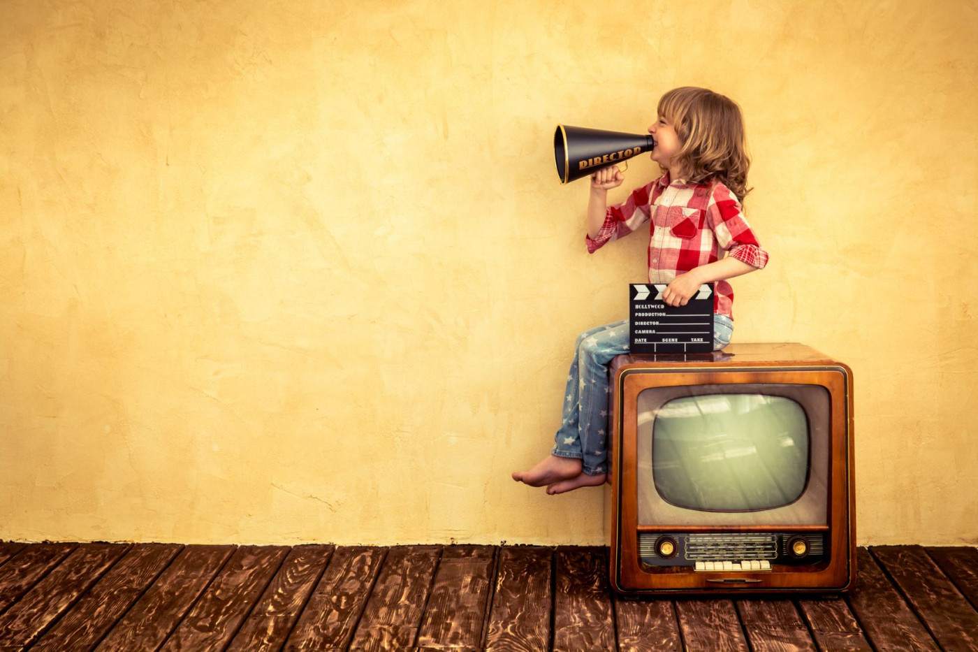 Ett barn sitter på en tjock-TV och ropar i en gammal megafon. I sin andra hand håller barnet i en filmklappa. 