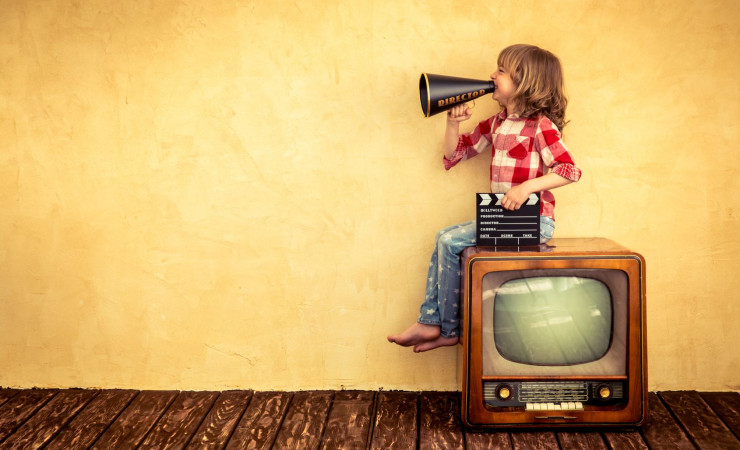 Ett barn sitter på en tjock-TV och ropar i en gammal megafon. I sin andra hand håller barnet i en filmklappa. 