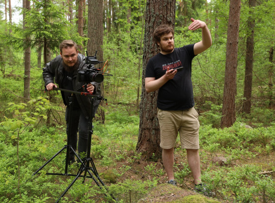 Filmset, med en filmfotograf som kikar ikameran. Framförkameran två ungdomar som sitter på marken i en skog. En av dem håller i ett skjutvapen.